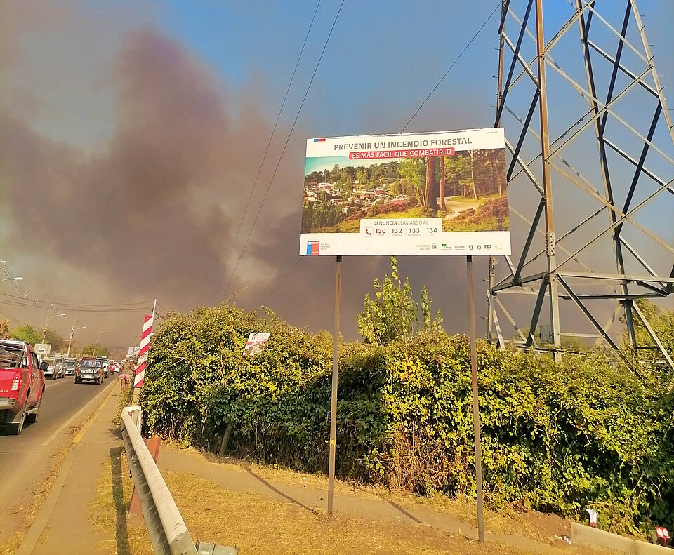 Letrero del Ministerio de Agricultura de Chile con la consigna "Prevenir un incendio forestal es mas fácil que combatirlo", mientras de fondo ocurre un…